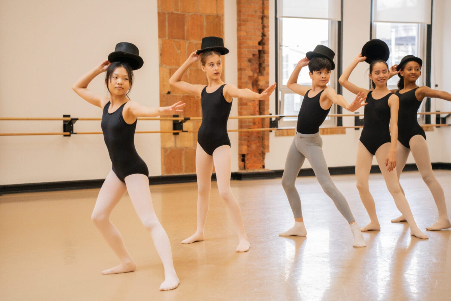 A group of students in a dance studio take Jazz class, lunging to the side and holding top hats above their heads.