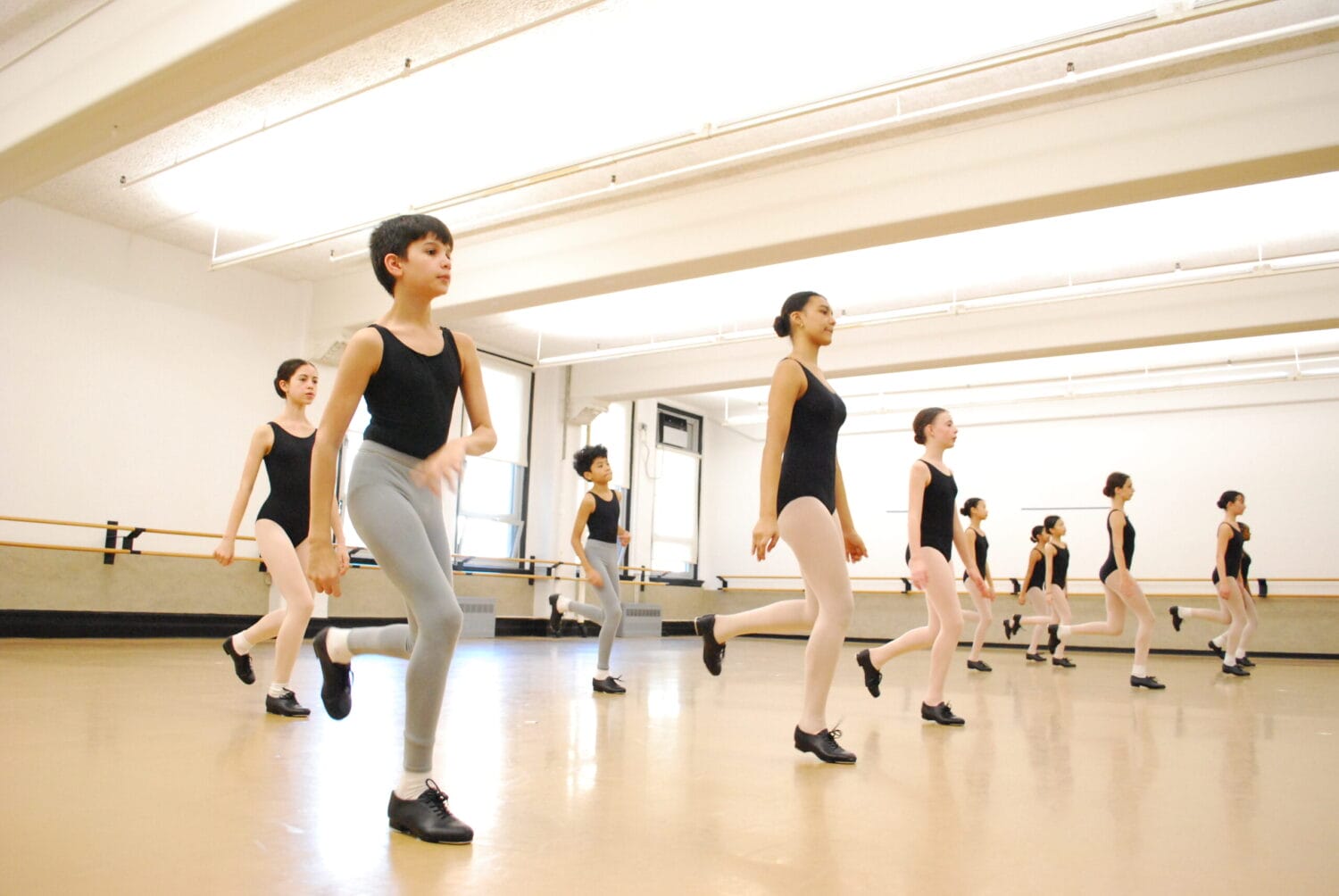 A group of dancers in a studio take a tap class, hopping on one foot with the other bent in the air.